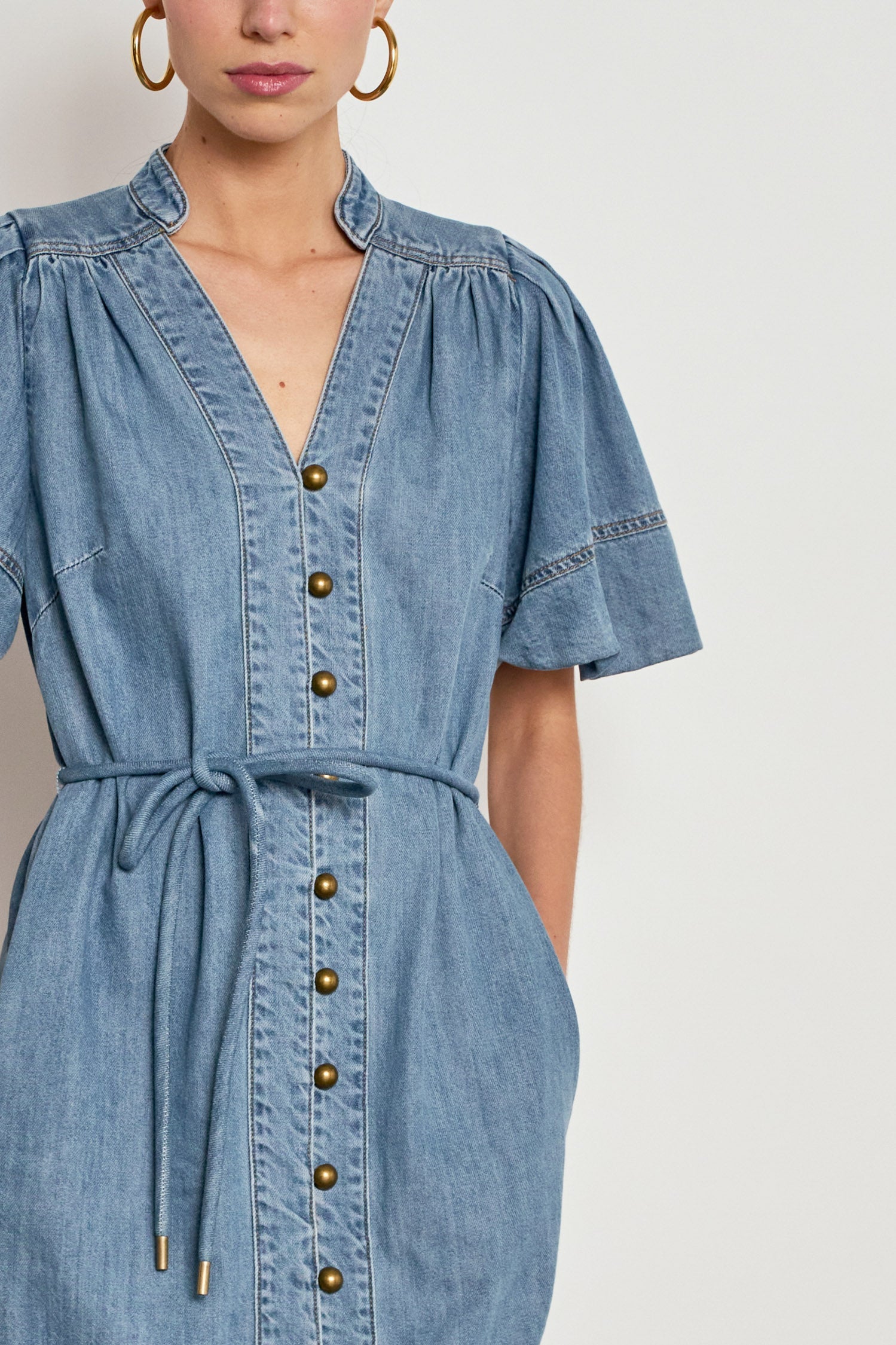 A close-up of a model wearing a light blue denim dress with a V-neck, button-down front, puff sleeves, and a matching tie belt at the waist.