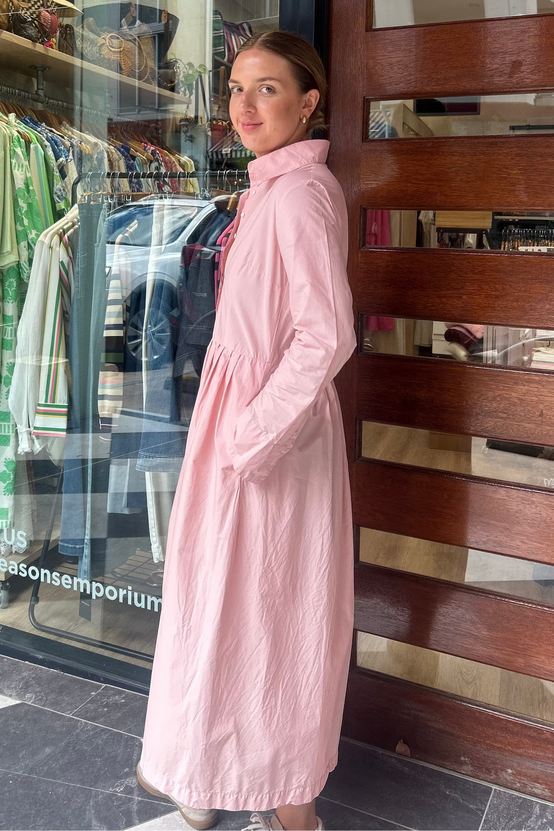 Woman in a pink coat standing outside a store with a glass door displaying clothing.