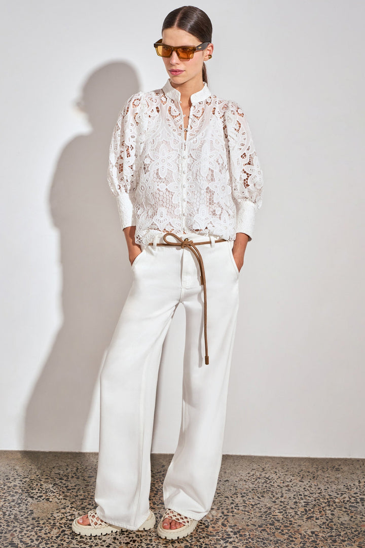 A model stands against a light gray wall wearing a white lace long-sleeve blouse, white wide-leg trousers with a thin brown tie belt, and beige sandals.