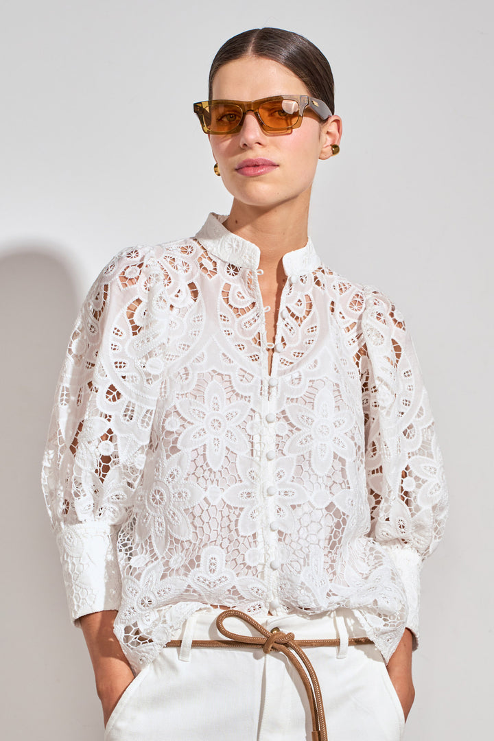 A model wearing a white long-sleeve lace blouse with a high collar, paired with white trousers and brown sunglasses, against a light gray studio background.