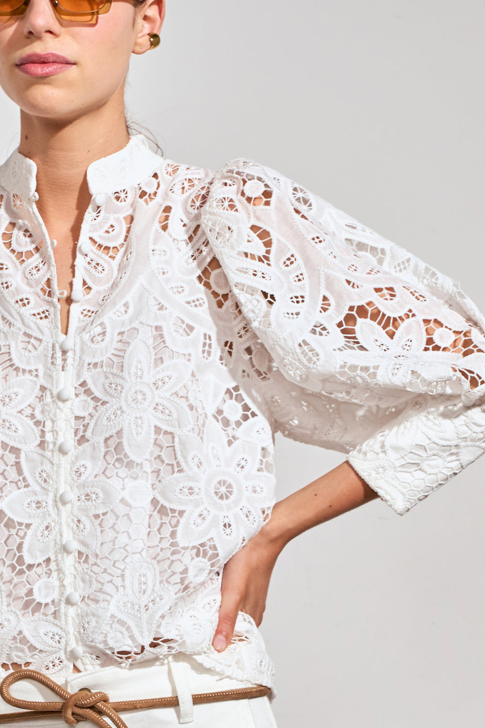 A close-up of a model wearing a white floral lace blouse with voluminous sleeves and a high collar, paired with a tan belt against a light gray background.