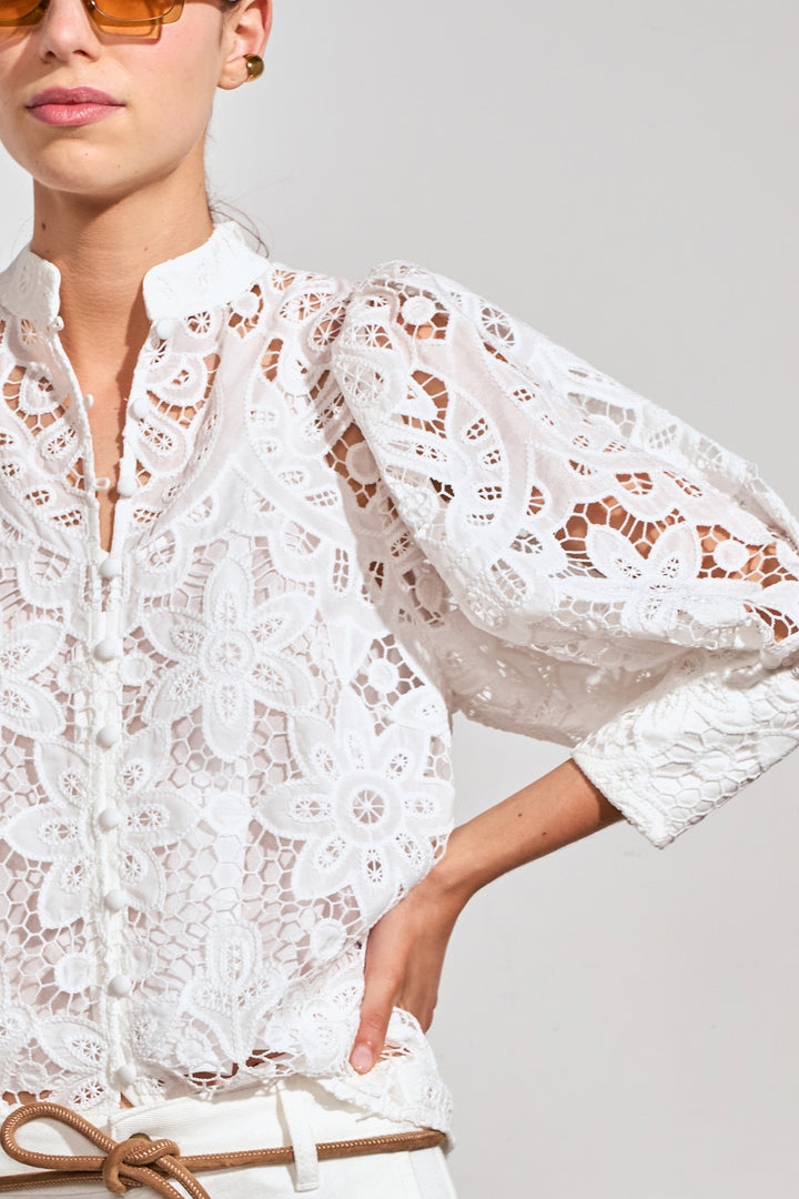 A close-up of a model wearing a white floral lace blouse with voluminous sleeves and a high collar, paired with a tan belt against a light gray background.