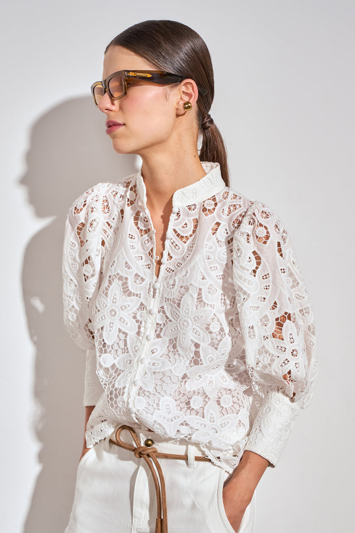 A woman wearing a white long-sleeve lace blouse with sheer puff sleeves and a stand collar, paired with white trousers and tortoiseshell glasses, set against a light gray wall with a soft shadow.