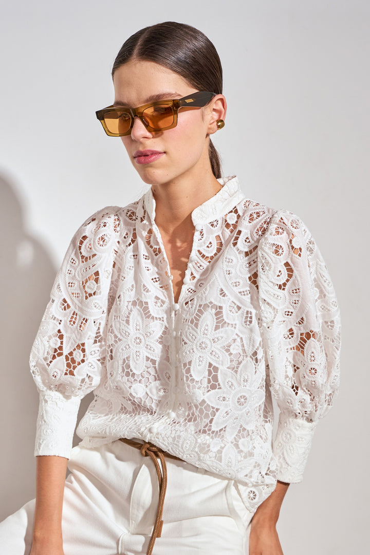A woman wearing a white floral lace blouse with puff sleeves and white pants, styled with amber-tinted sunglasses against a light gray background.