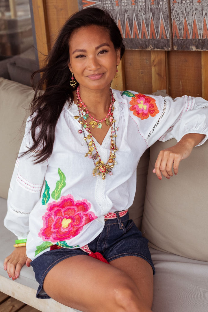 A woman sitting on a beige outdoor sofa wearing a white long-sleeve blouse with vibrant pink and orange floral embroidery, dark denim shorts, and layered gold necklaces.