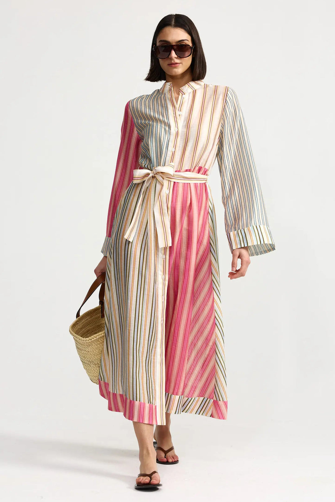 Woman wearing a long, multicolored striped dress holding a straw bag on a white background. Aston Studio Neotta Dress Peony Multi Stripe