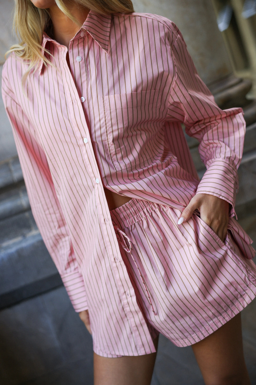 A woman wearing a pink and white vertically striped button-down shirt and matching shorts, photographed in a real architectural setting with stone steps in the background.