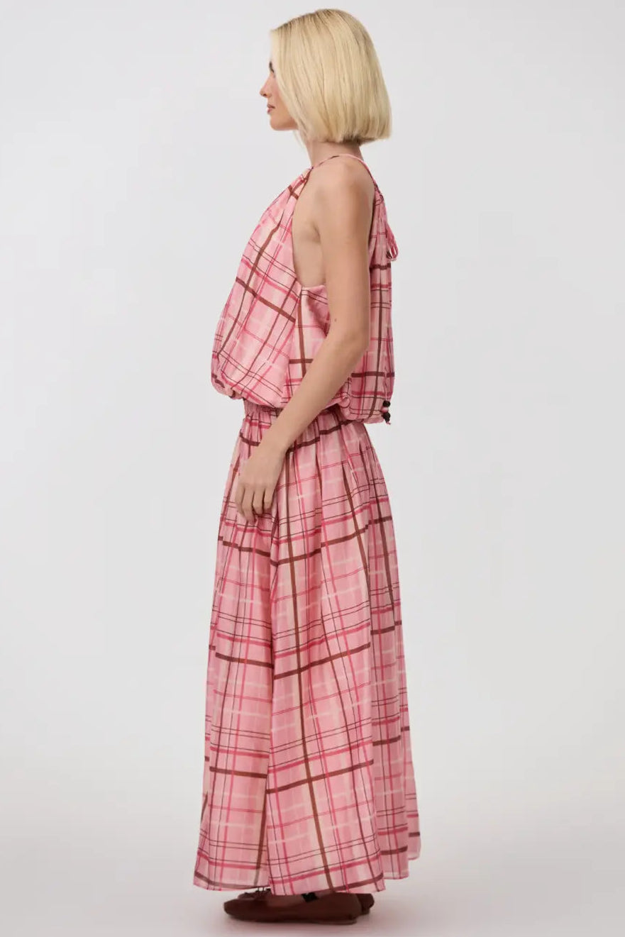 A woman with a short blonde bob is shown from the side profile wearing a pink and red plaid halter-neck maxi dress with a matching belt and skirt, standing against a plain light gray studio background.