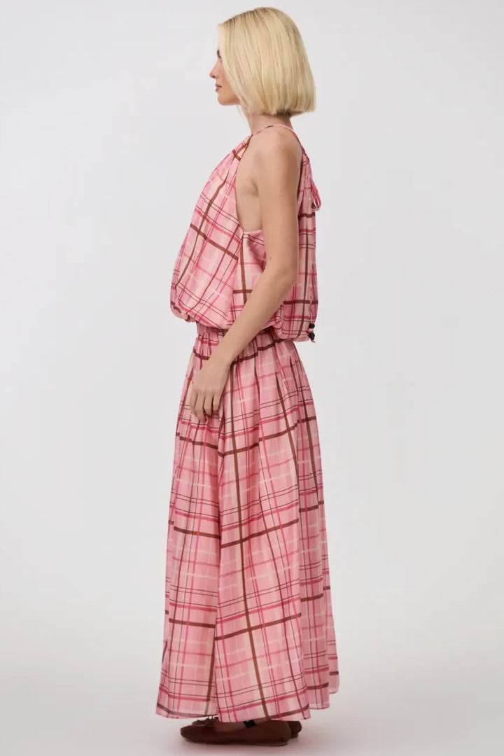 A woman with a short blonde bob is shown from the side profile wearing a pink and red plaid halter-neck maxi dress with a matching belt and skirt, standing against a plain light gray studio background.