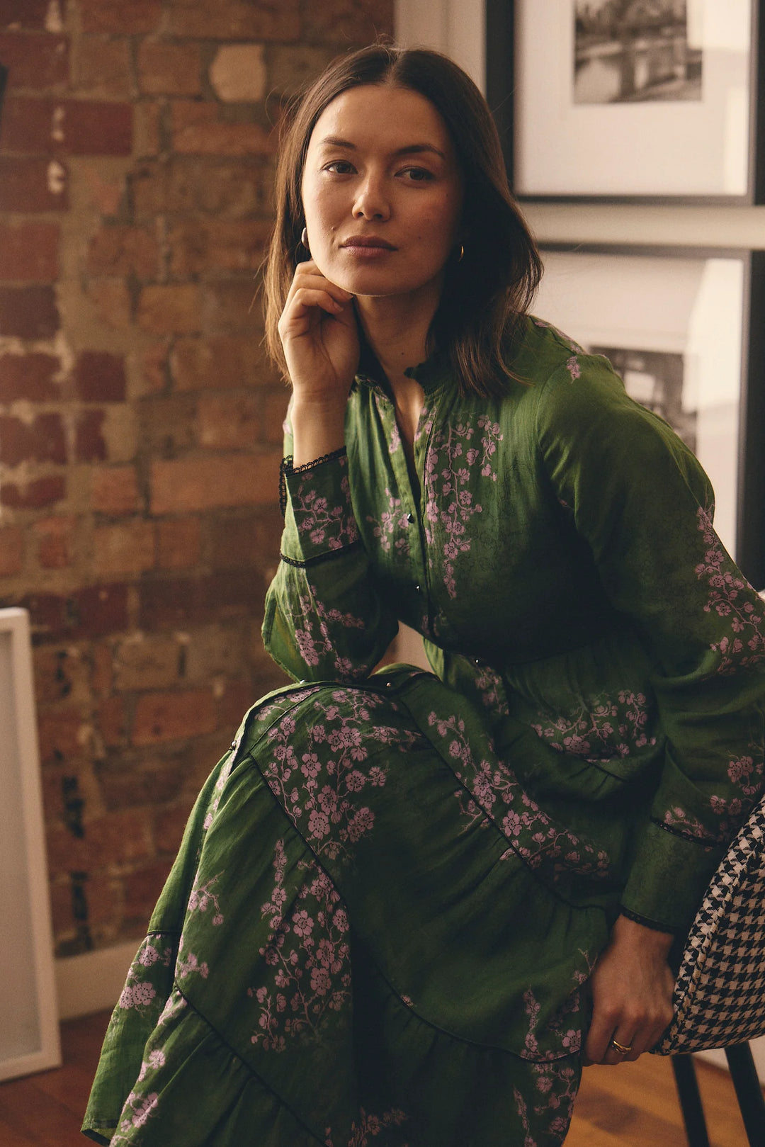 Woman in a green floral outfit sitting against a brick wall.
