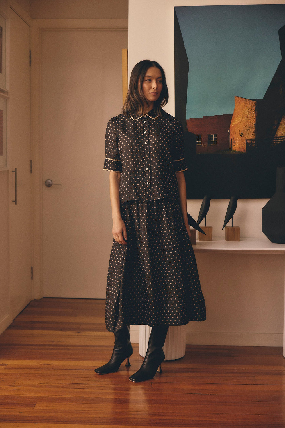 Woman in a black patterned dress standing in a room with a painting on the wall.