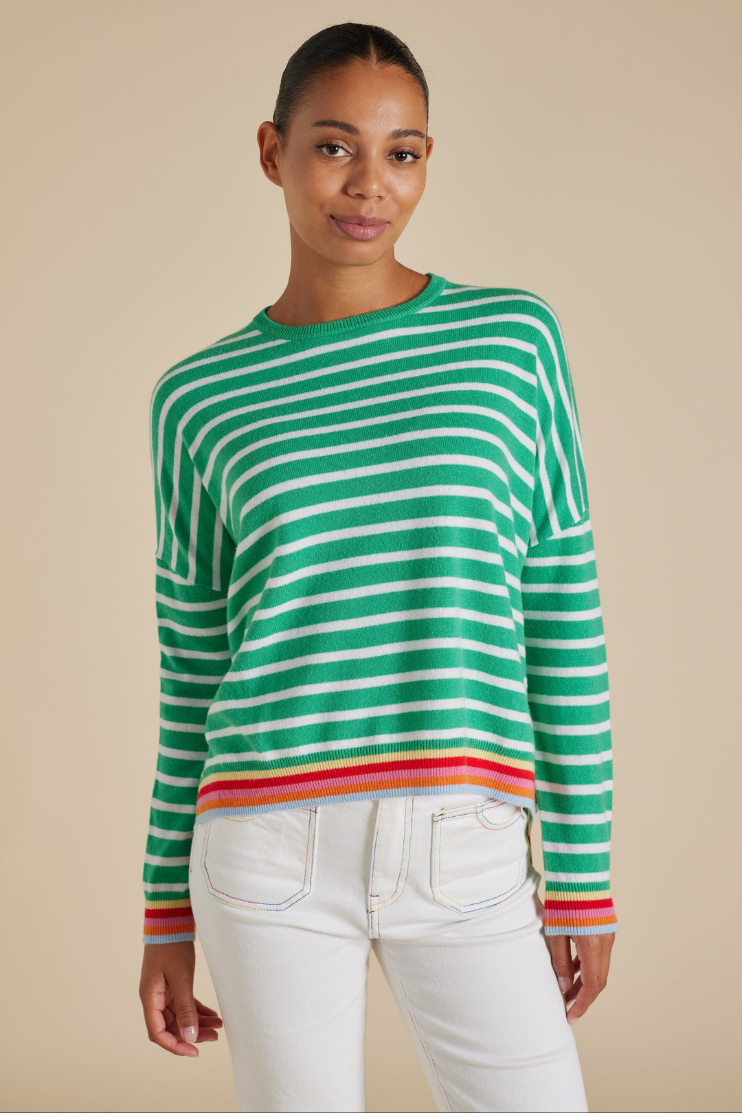 A woman wearing a pine green and white striped sweater with contrast rainbow-striped cuffs and hem, paired with white pants.