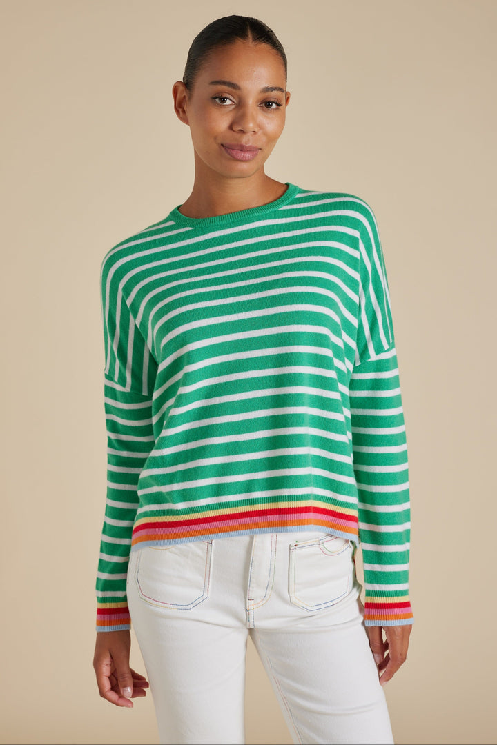 A woman wearing a pine green and white striped sweater with contrast rainbow-striped cuffs and hem, paired with white pants.