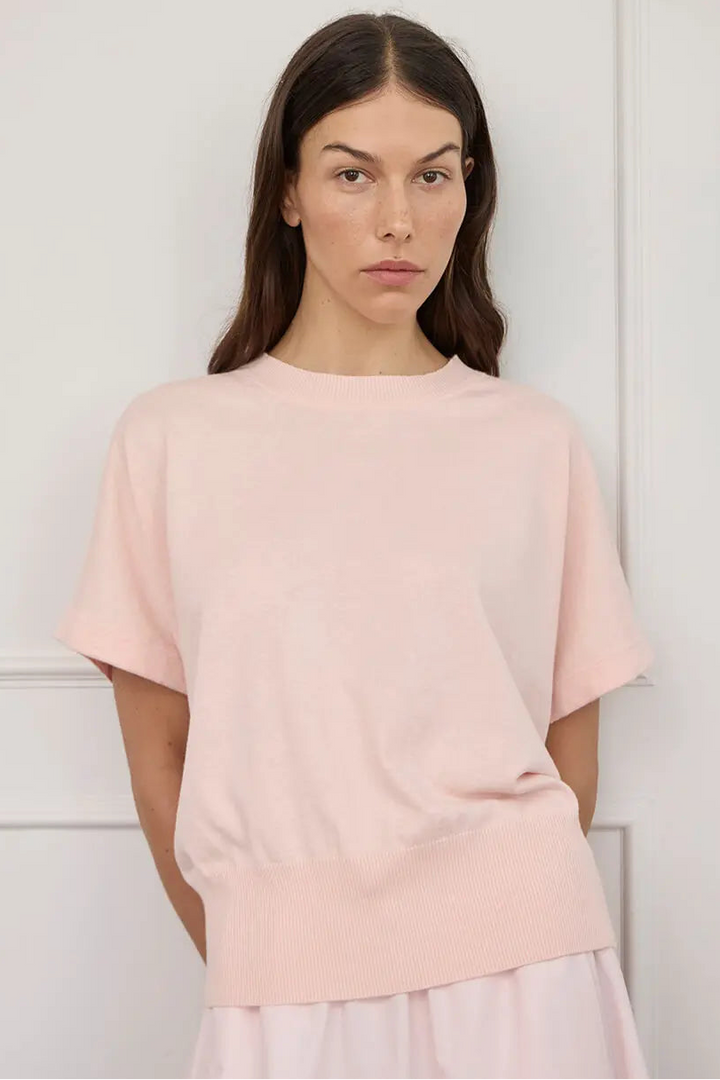A woman wearing a pale pink short-sleeve knit top with a wide ribbed hem, standing against a white paneled wall.