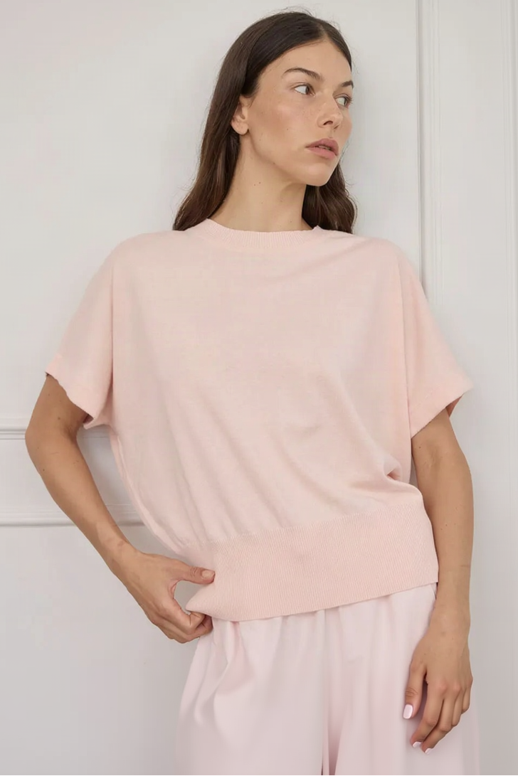 A woman wearing a light pink short-sleeve knit top with a wide ribbed hem, standing against a white paneled wall.