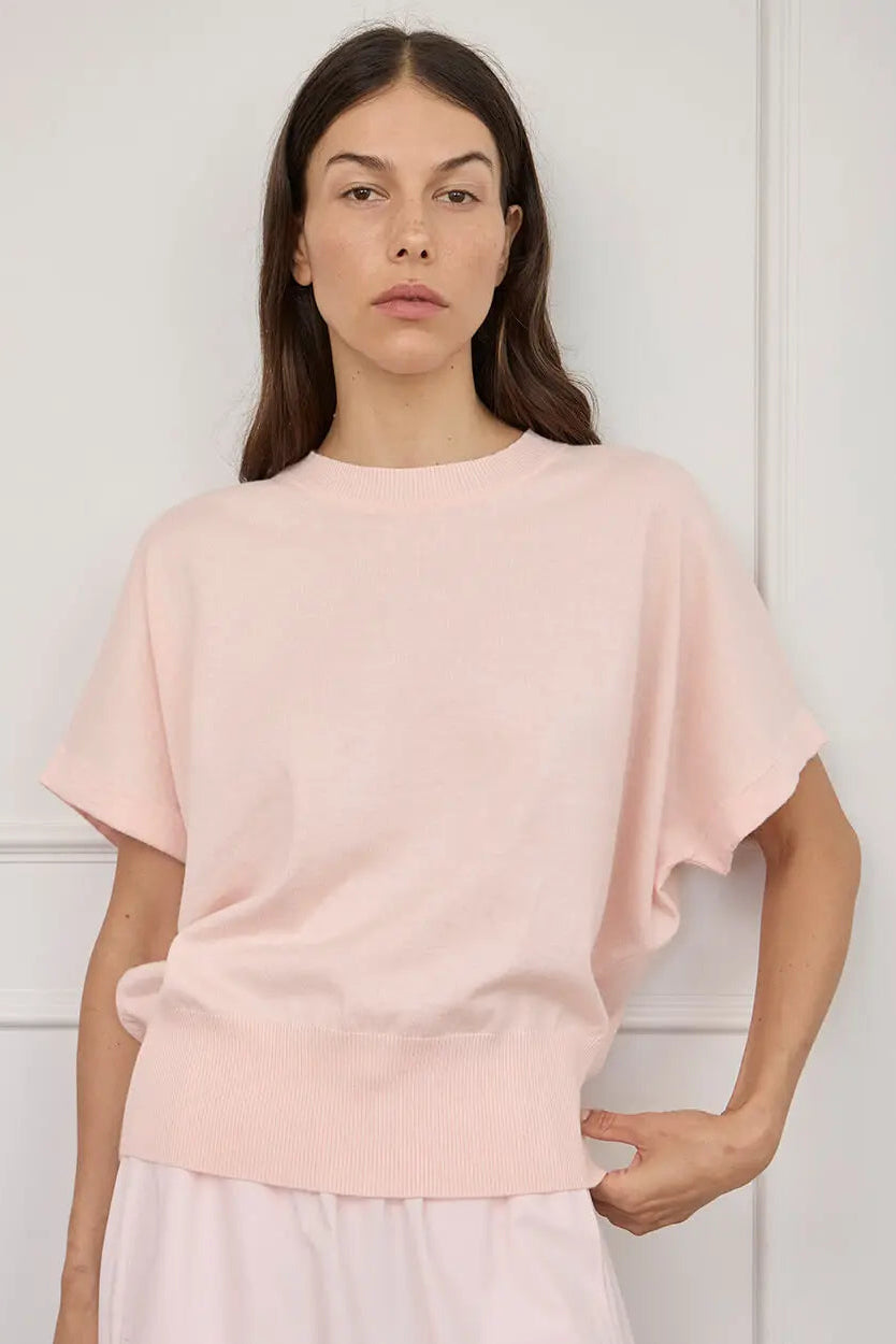 A woman modeling a light pink short-sleeve knit top with a relaxed, oversized fit and a ribbed hem, paired with a matching pale pink skirt against a white paneled wall.