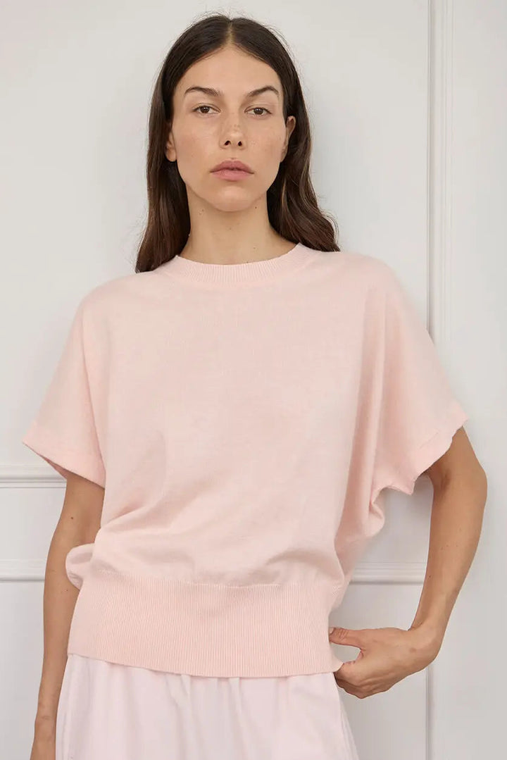 A woman modeling a light pink short-sleeve knit top with a relaxed, oversized fit and a ribbed hem, paired with a matching pale pink skirt against a white paneled wall.
