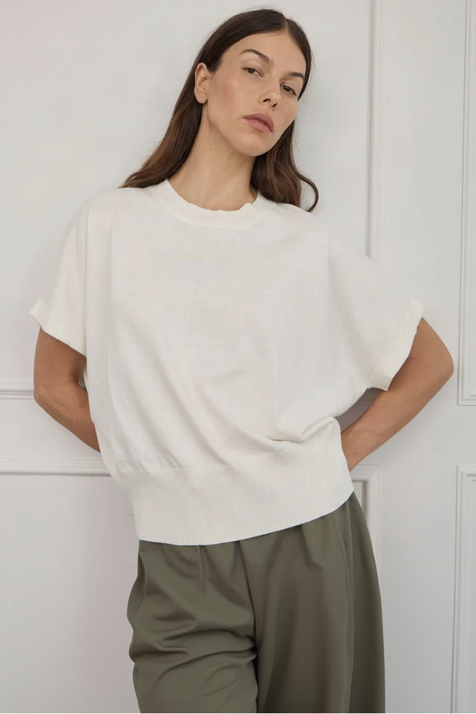 A woman wearing a loose-fitting, short-sleeve crewneck knit top in off-white, paired with olive green bottoms, standing against a white paneled wall.