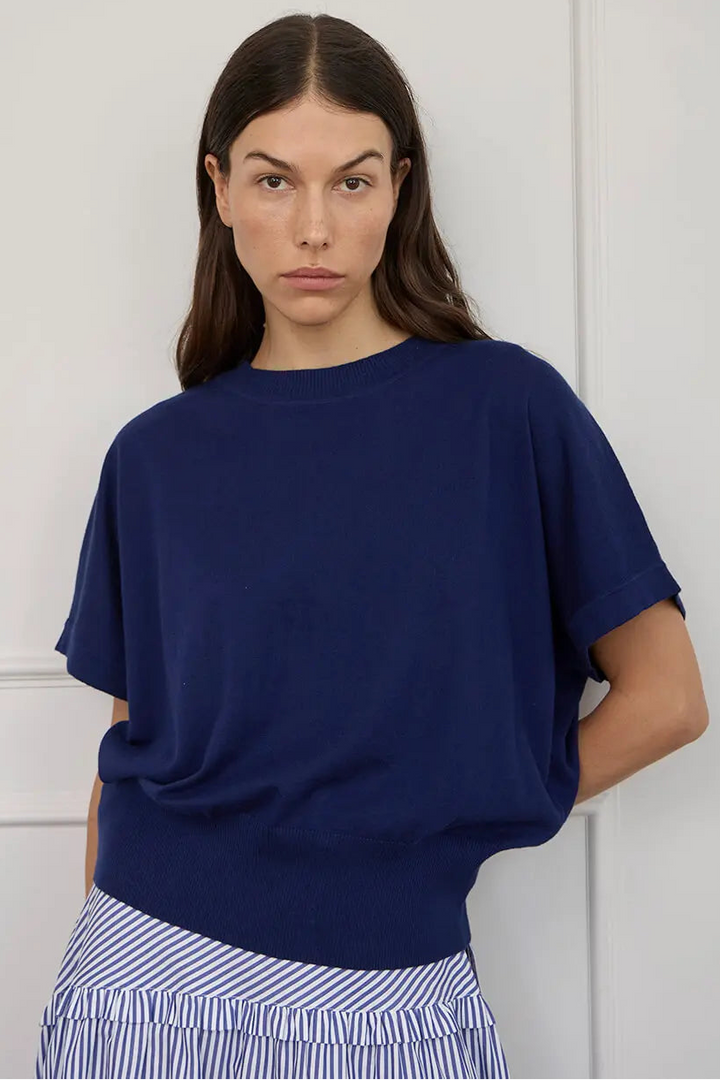 A woman wearing a loose, short-sleeve navy blue knit top and blue-and-white striped bottoms, standing against a white paneled wall.