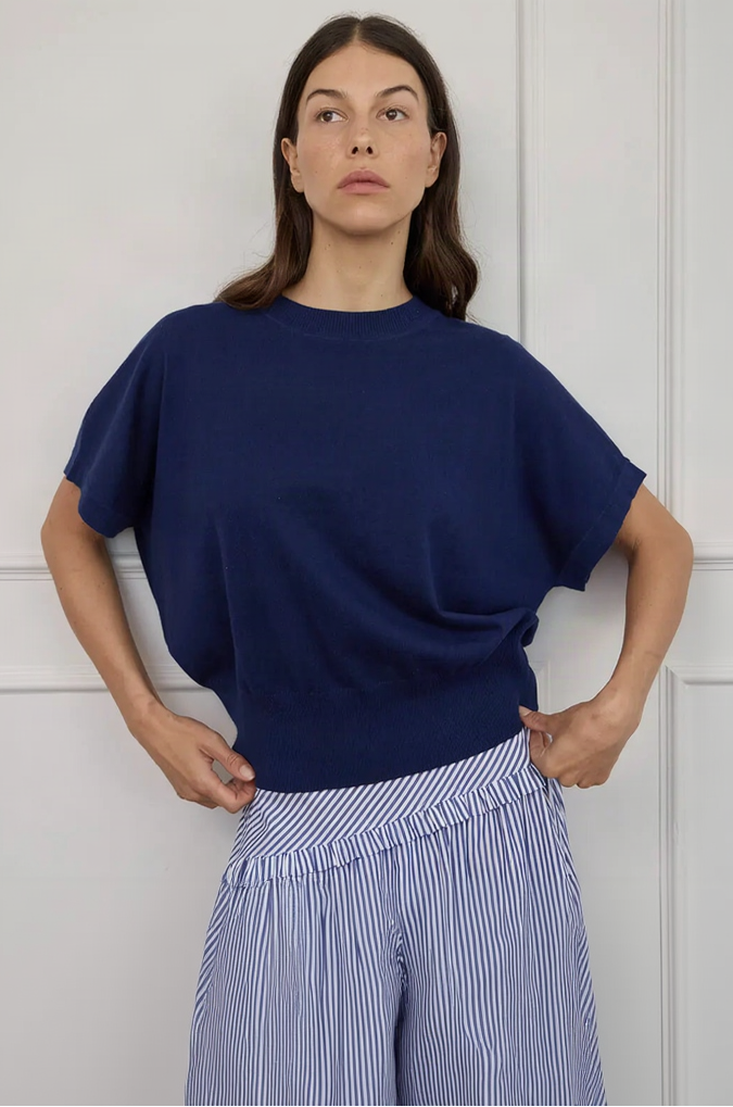 A woman wearing a navy blue short-sleeve knit top with a relaxed, oversized fit, paired with blue and white striped bottoms, posing against a white paneled wall.