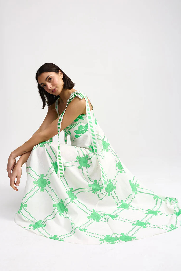 Woman wearing a white dress with green floral patterns on a white. Aston Studio Pippa Dress Mediterranean Fernbackground