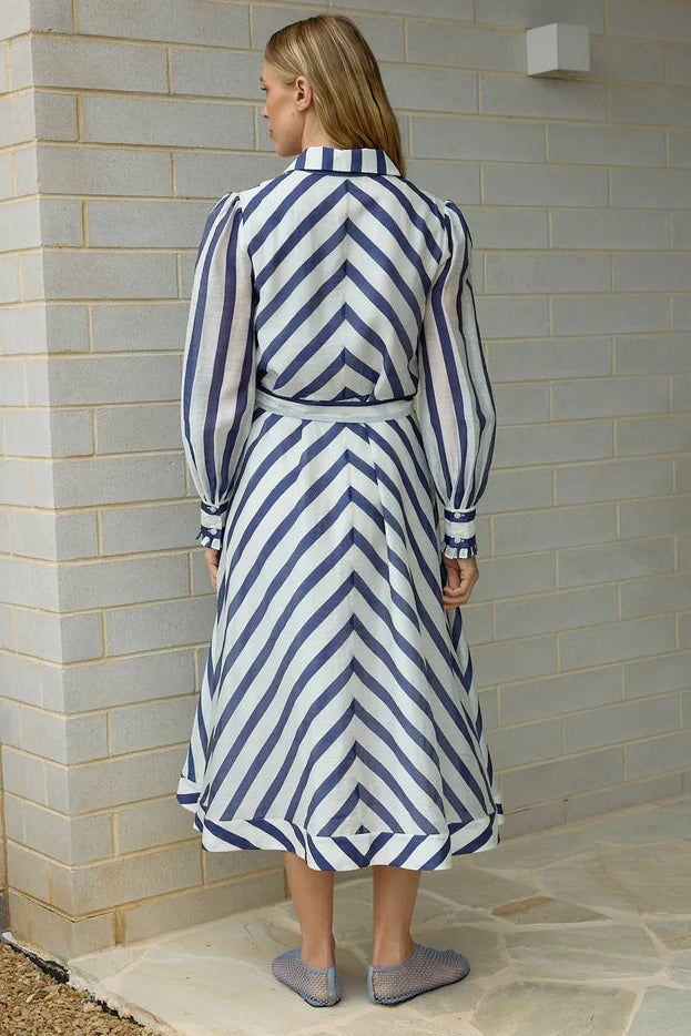 Woman wearing a blue and white striped dress standing against a light brick wall.