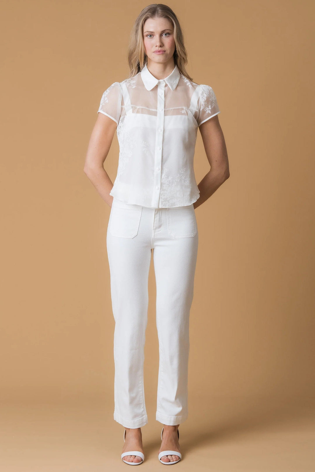 A woman modeling a white short-sleeve blouse with sheer mesh sleeves and white straight-leg pants, standing against a solid tan studio backdrop.
