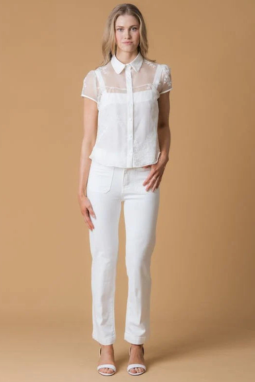 A woman modeling a white short-sleeve blouse with sheer mesh sleeves and white pants, standing against a solid tan studio background.