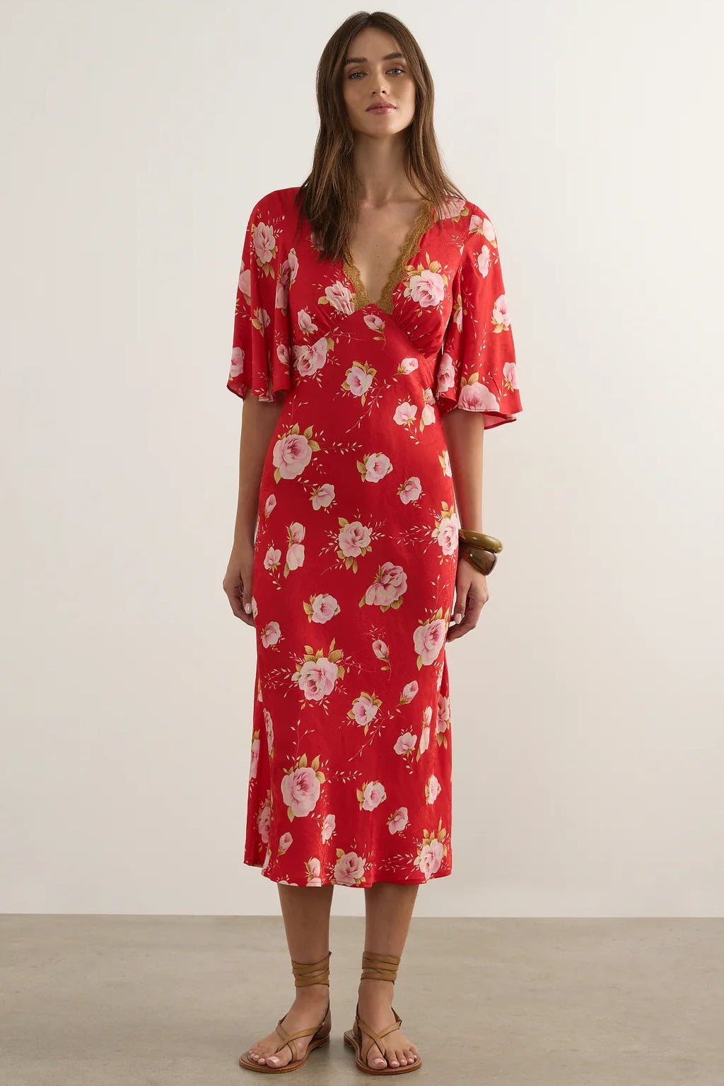 A woman modeling a red midi dress with a white and pink floral print, featuring a wrap-style V-neckline and short flutter sleeves, set against a plain light gray studio background.