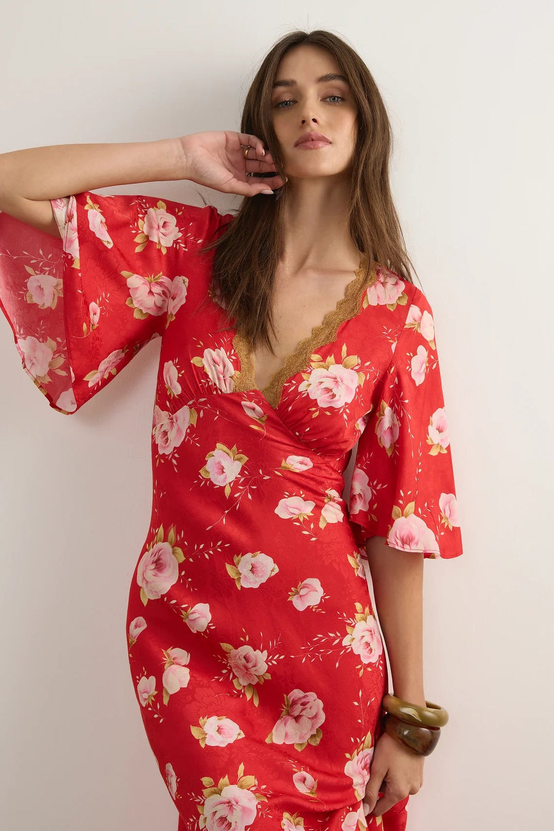 A woman modeling a red wrap-style dress with a pink and white floral print, featuring wide short sleeves and a lace-trimmed V-neckline, posed against a plain off-white wall.