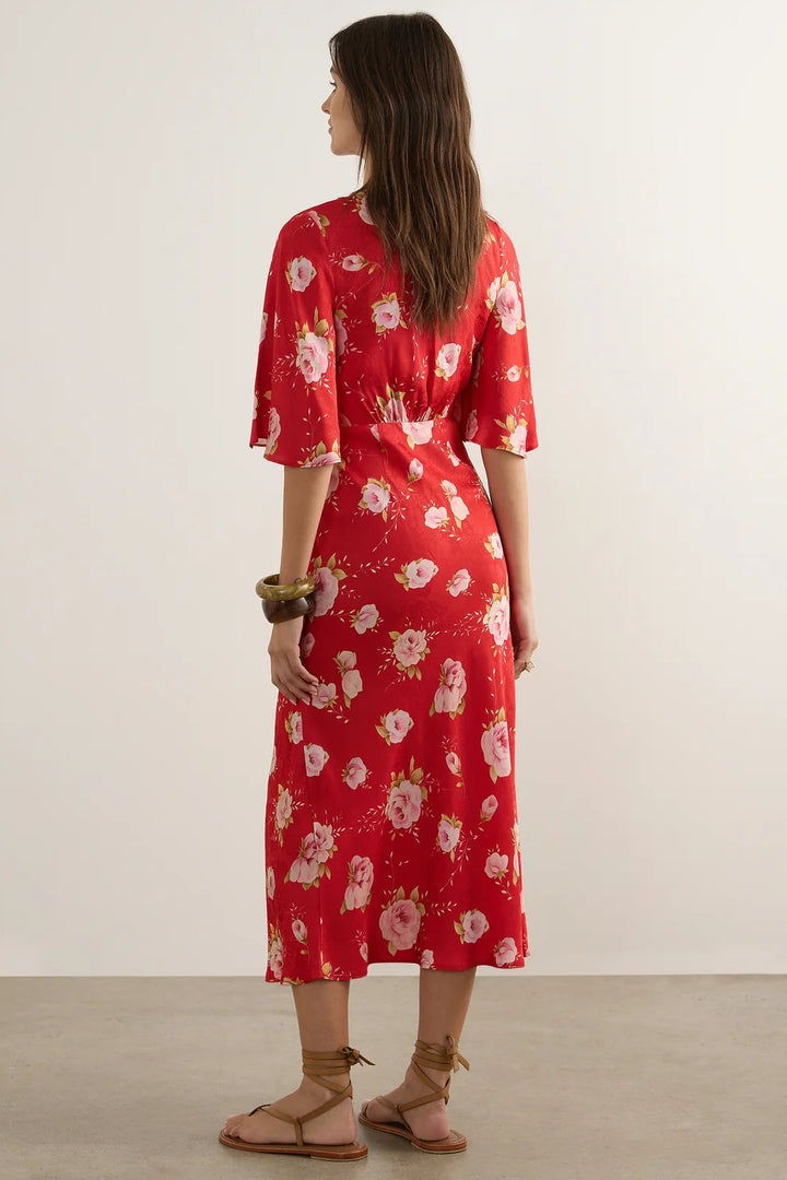 A woman stands with her back to the camera wearing a red midi dress with a white and pink floral pattern, paired with brown leather sandals, in a minimalist studio setting.