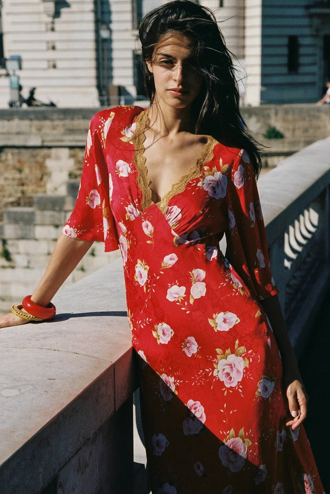 A woman wearing a red floral maxi dress with gold lace trim, leaning on a stone railing in an outdoor setting.