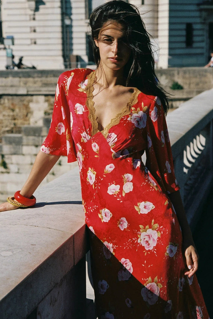 A woman wearing a red floral maxi dress with gold lace trim, leaning on a stone railing in an outdoor setting.
