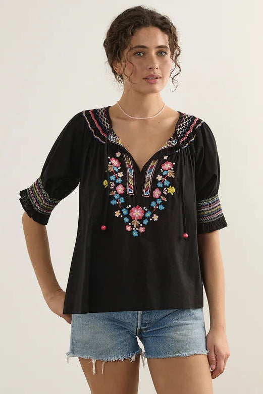 A woman modeling a black short-sleeve top with colorful floral embroidery on the chest and sleeve cuffs, paired with denim shorts and white slide sandals, standing against a plain light gray studio background.