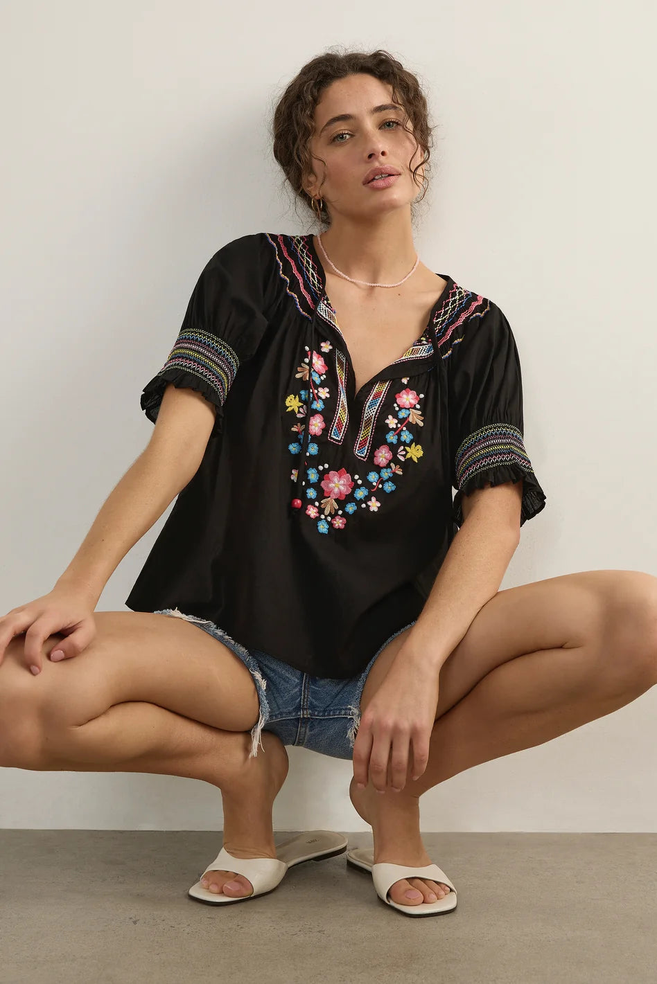 A woman crouching in denim shorts and white slide sandals, wearing a black short-sleeve bohemian-style blouse with colorful floral embroidery and striped trim on the neckline and cuffs, set against a plain light gray wall.