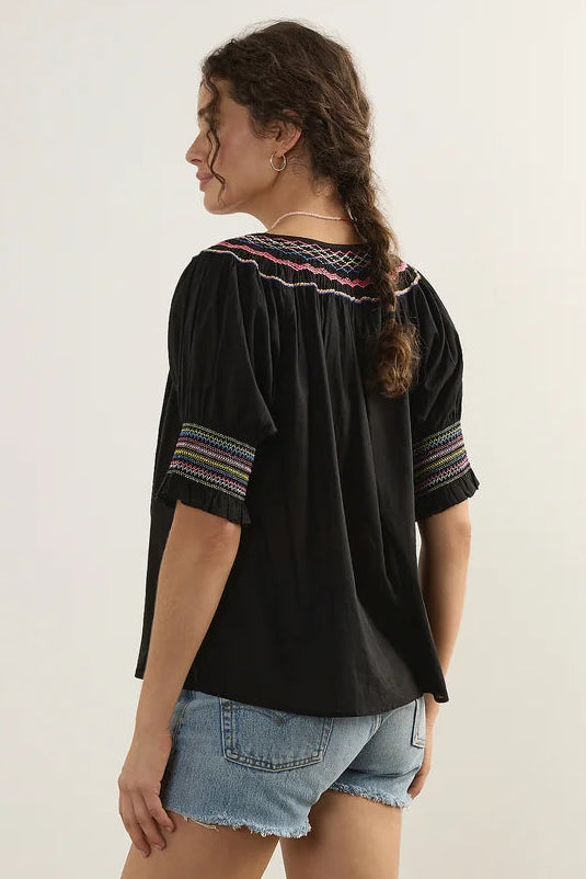 A woman modeling a black short-sleeve blouse with colorful striped trim at the neckline and sleeve cuffs, paired with denim shorts and white slide sandals, photographed in a studio setting.