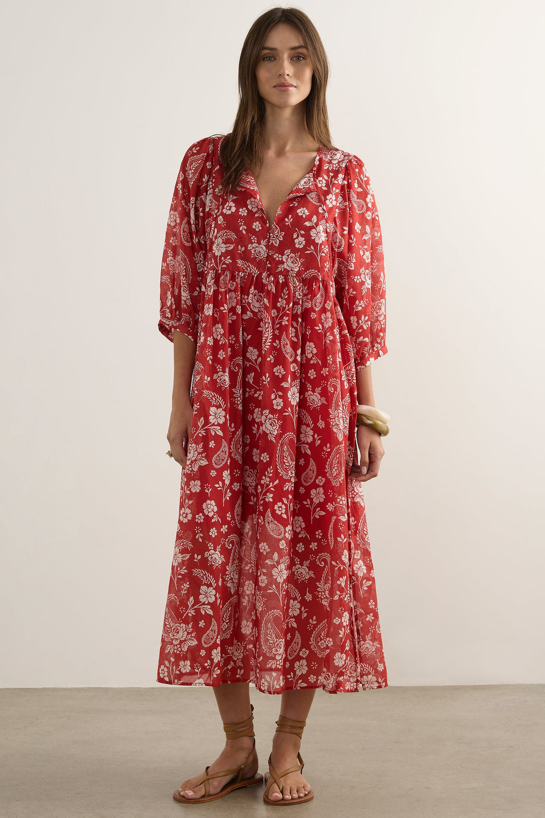 A woman modeling a red midi dress with a white floral paisley pattern, standing against a plain off-white studio background.