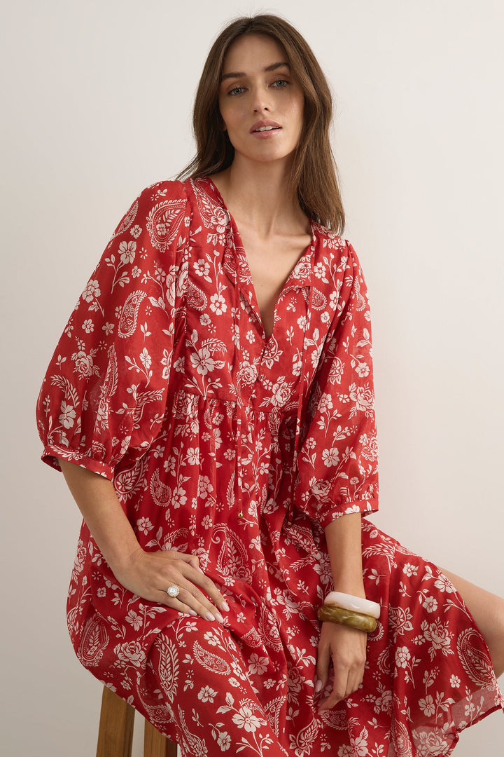 A woman wearing a red and white paisley print dress, sitting on a stool against a plain off-white background.