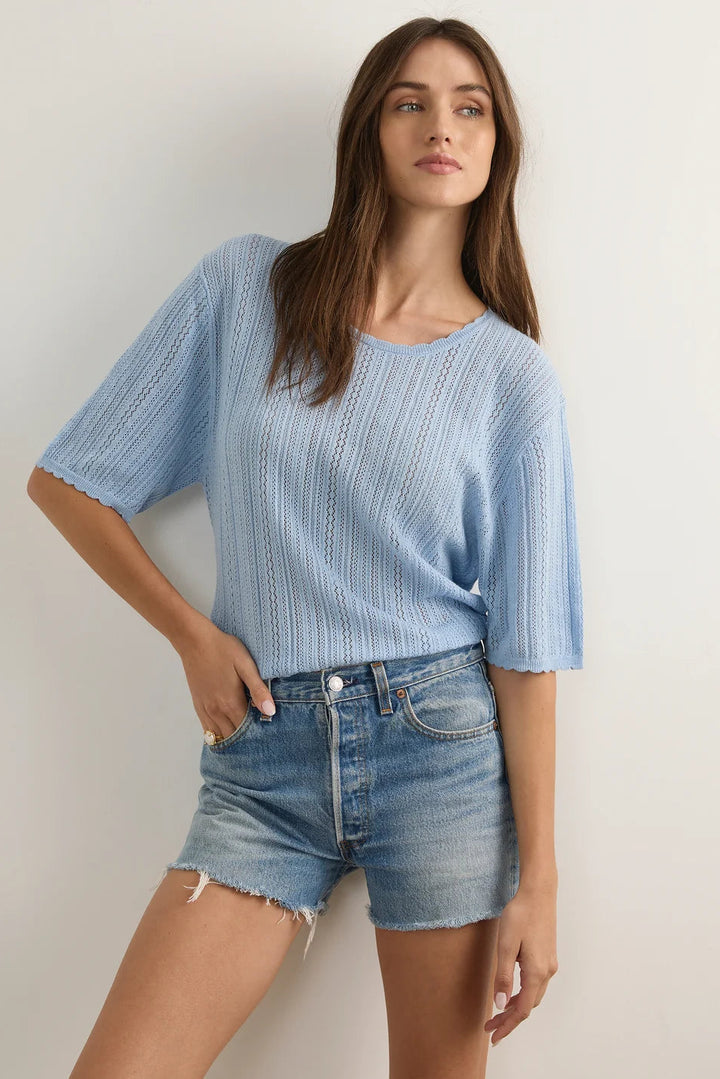 A woman modeling a light blue short-sleeve knit top with a textured openwork pattern and frayed edges, paired with denim shorts, against a plain off-white wall.
