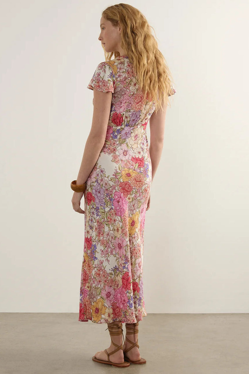 A woman stands with her back to the camera wearing a short-sleeve, knee-length floral dress with a fitted silhouette and a multicolor print on a light background. She is wearing brown strappy sandals against a plain off-white studio wall and concrete floor.