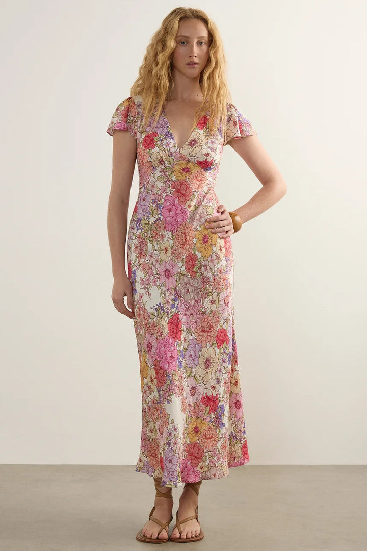 A woman modeling a short-sleeve, V-neck maxi dress with a dense pink, red, yellow, and purple floral pattern on a light cream background, set against a neutral studio backdrop.