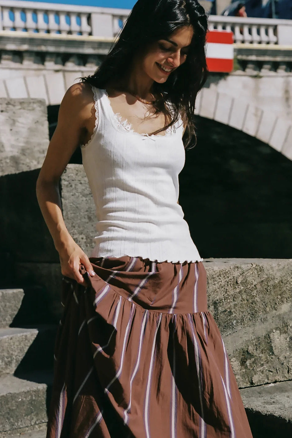 A woman wearing a white ribbed knit tank top and a brown tiered skirt with thin white stripes, standing on stone steps outdoors.
