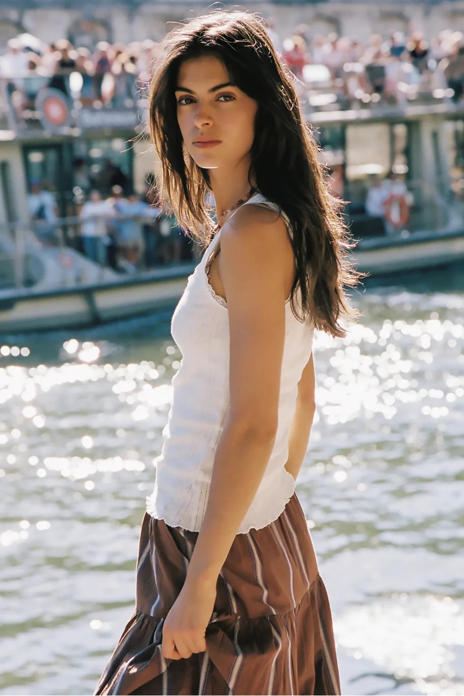 A woman standing outdoors by a body of water with boats and a crowd in the background, wearing a white sleeveless top and a brown skirt with thin light stripes.