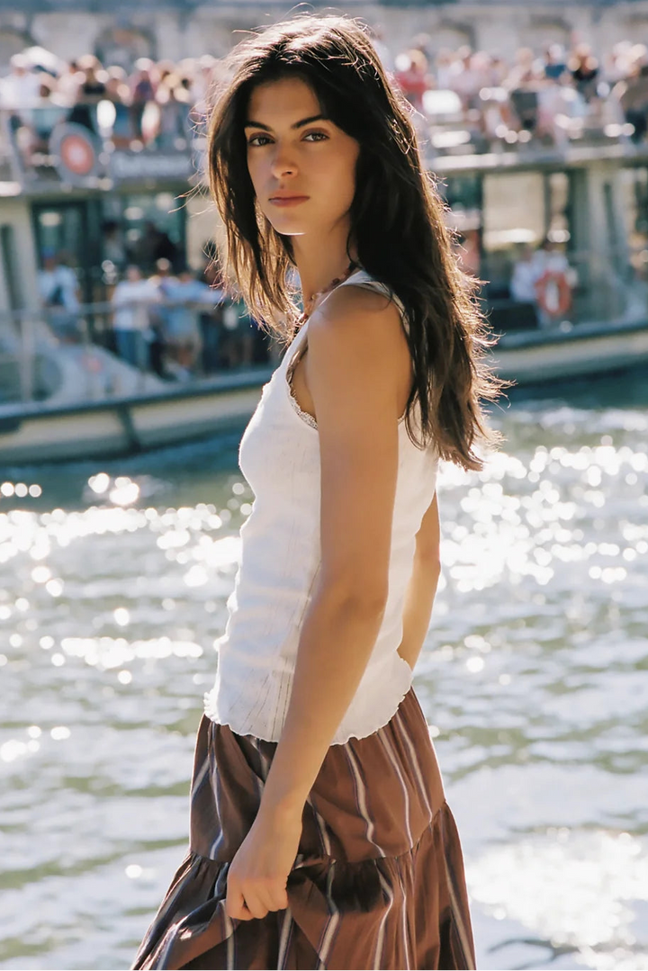 A woman standing outdoors by a body of water with boats and a crowd in the background, wearing a white sleeveless top and a brown skirt with thin light stripes.