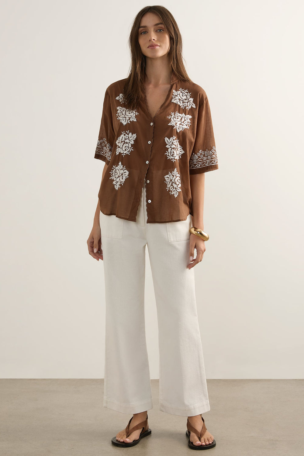 Woman wearing a brown floral shirt and white pants against a plain background