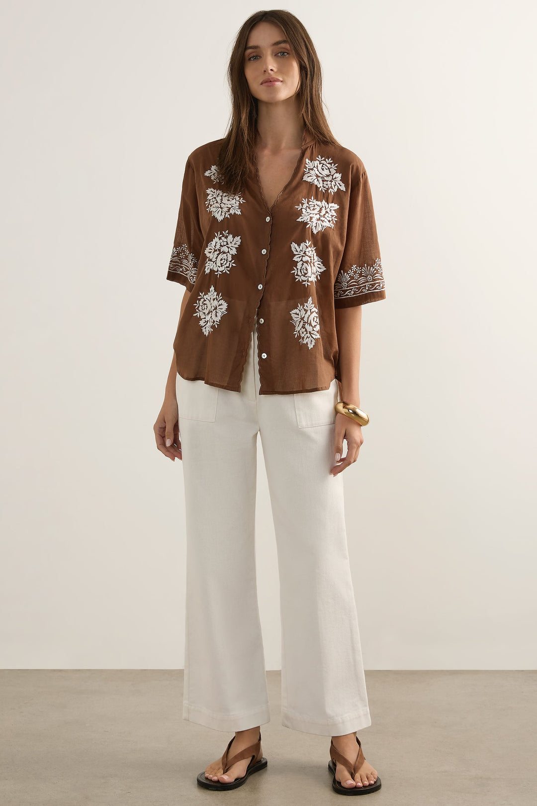 Woman wearing a brown floral shirt and white pants against a plain background