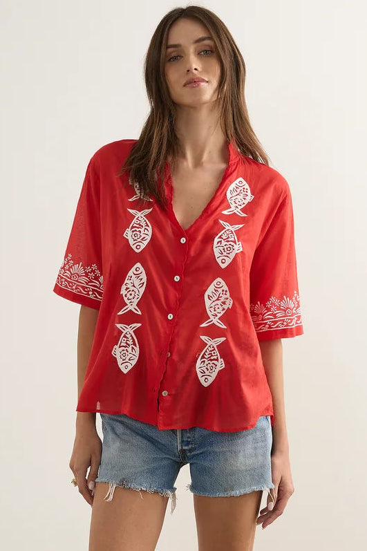 A woman modeling a red short-sleeve button-up shirt with white embroidered fish motifs, paired with denim shorts and brown sandals, standing against a plain light beige studio wall.