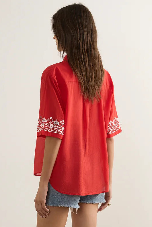 A woman is shown from behind wearing a bright red short-sleeve blouse with white floral embroidery on the cuffs, paired with denim shorts and brown strappy sandals, standing against a neutral studio background.