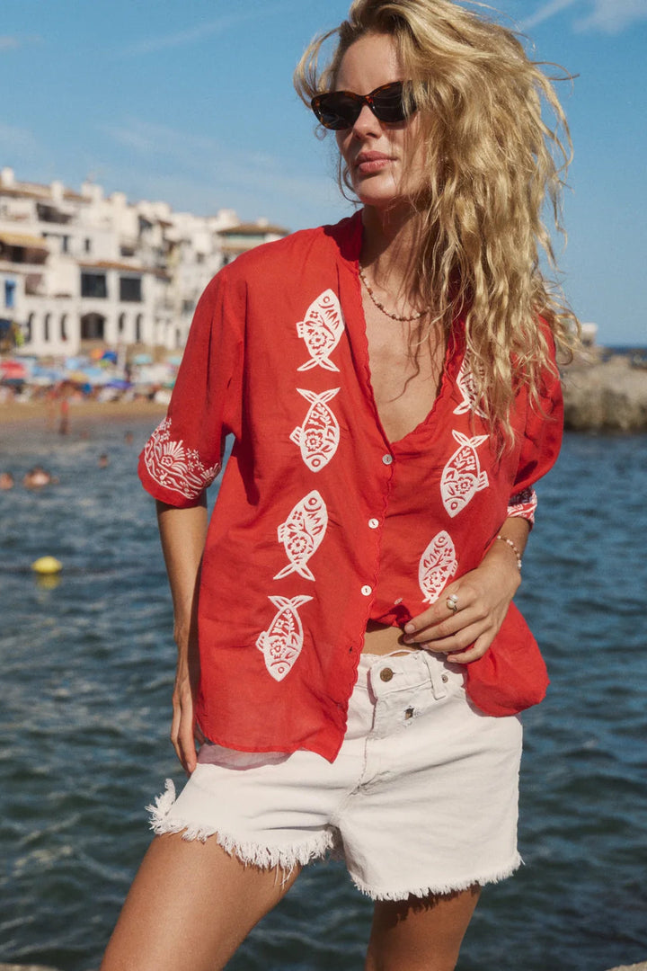 A woman wearing a red short-sleeve button-up shirt with white fish prints and white denim shorts, standing by the sea with beach buildings in the background.
