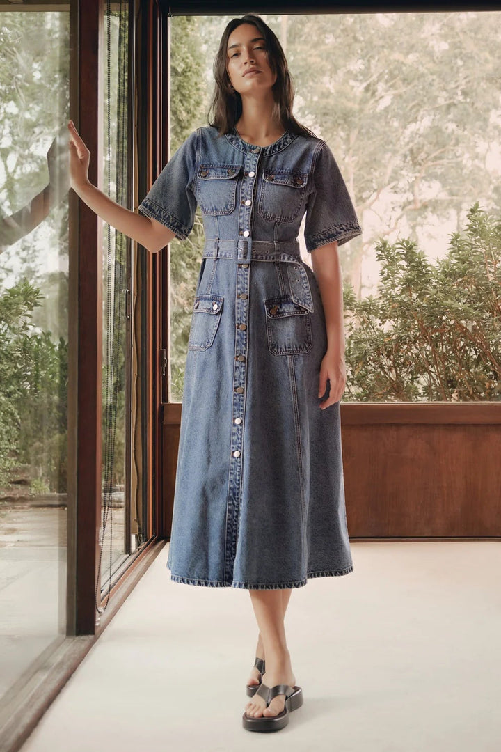 Woman wearing a denim dress standing in a room with large windows showing greenery outside.